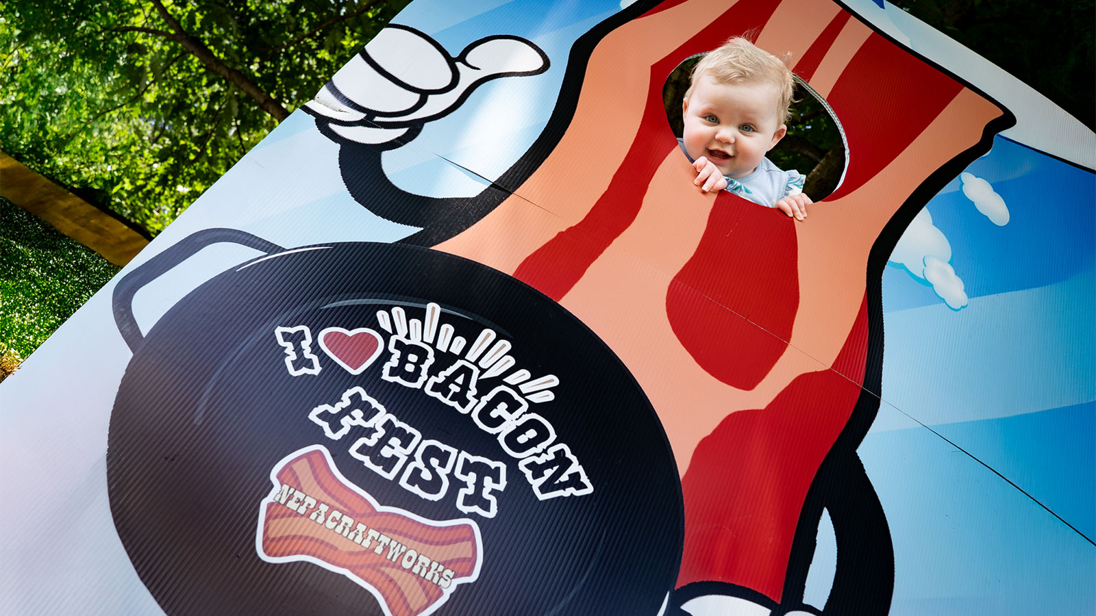 Baby showing her face through the opening in a photo cutout board depicting a strip of bacon at I Love Bacon Fest in Forty Fort, PA.