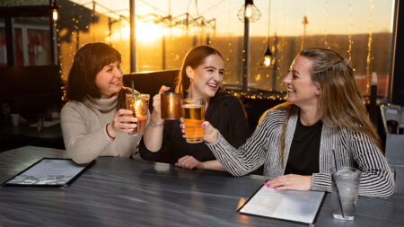 Three women cheersing and smiling with a sunset in the background at River Grill in Plains, PA.