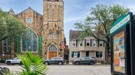 A stone church with a tall bell tower stands beside a residential building in downtown Stroudsburg, PA, with parked cars and a sign reading 