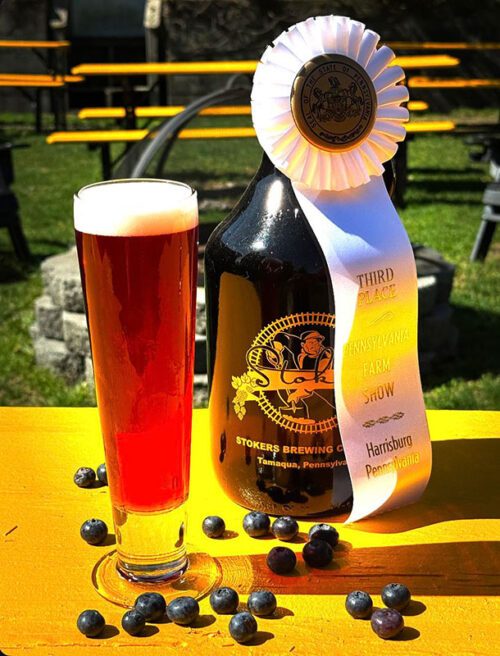 A dark, red beer in a tall pilsner glass with blueberries and a growler bottle and an award ribbon next to it at Stoker's Brewing Co. in Tamaqua, PA.