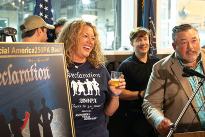 A woman smiles while holding a glass of beer next to a poster for Declaration Lager