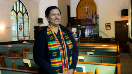 A woman stands inside a church adorned with colorful stained-glass windows.