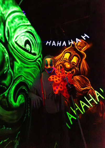 An actor dressed as a clown stands in a room with paintings of laughing clowns in neon colors at Reaper's Revenge in Scott Twp., PA near Scranton.
