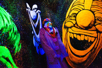 An actor dressed as a clown stands in a room with paintings of laughing clowns in neon colors at Reaper's Revenge in Scott Twp., PA near Scranton.