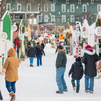 Visitors at Christmas Market at Christmas in Montrose, PA, walk through a snowy outdoor Christmas market lined with decorated vendor huts and festive lights.