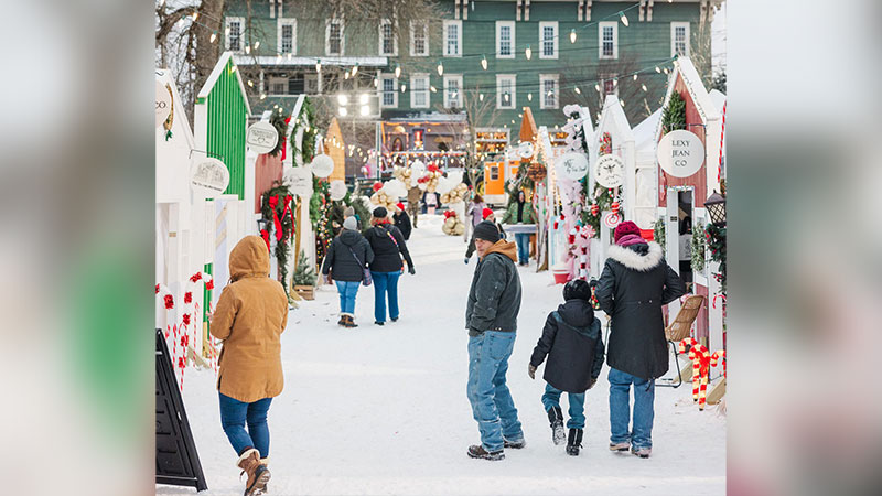 Visitors at Christmas Market at Christmas in Montrose, PA, walk through a snowy outdoor Christmas market lined with decorated vendor huts and festive lights.