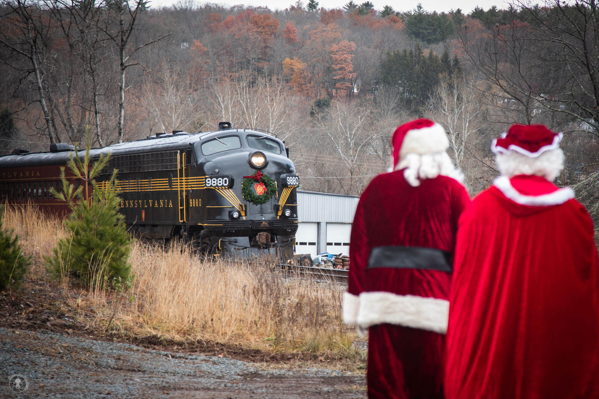 Santa and Mrs. Claus stand near train tracks watching an approaching vintage locomotive decorated with a Christmas wreath at the Santa Express at Stourbridge Line in Honesdale.