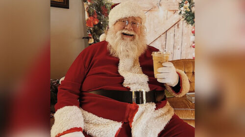 Santa Claus sits indoors with a drink in hand, smiling warmly beside rustic holiday décor at Santa at the Farmstand.