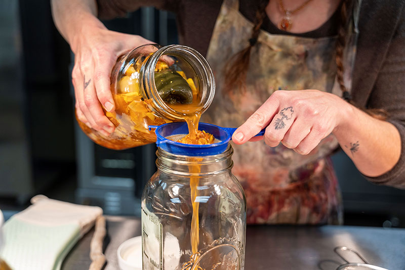 Storms End Homestead founder pouring infused herbal liquid through a strainer while producing elderberry syrup at the Hazleton Kitchen Incubator in Hazleton, PA.