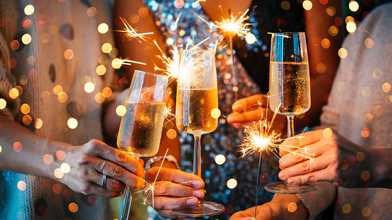 Close-up of hands holding champagne glasses and lit sparklers against festive golden bokeh lights.