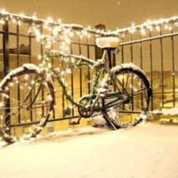 Photo of a bicycle covered in snow and wrapped in glowing holiday string lights on a railing overlooking a nighttime cityscape.