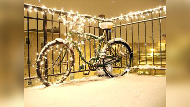 Photo of a bicycle covered in snow and wrapped in glowing holiday string lights on a railing overlooking a nighttime cityscape.