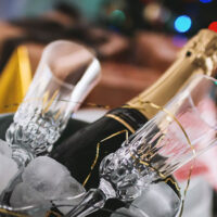 Bottle of champagne nestled in an ice bucket beside two crystal flutes with soft holiday lights in the background.