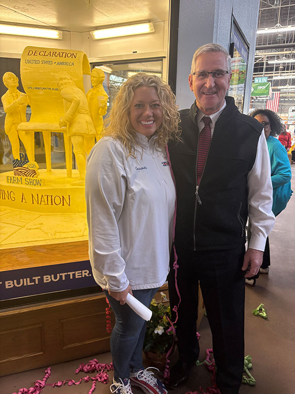 Two people posing for a photo in front of the butter sculpture at the PA Farm Show in Harrisburg, PA.