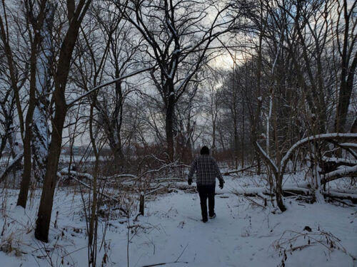 A lone hiker follows a snowy riverside trail through leafless trees at Kirby Park in Wilkes-Barre, PA, during winter.