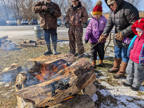 Families roast marshmallows over an open fire during Riverfront Parks Winterfest in Wilkes-Barre, PA, bundled up in winter coats on a chilly day.