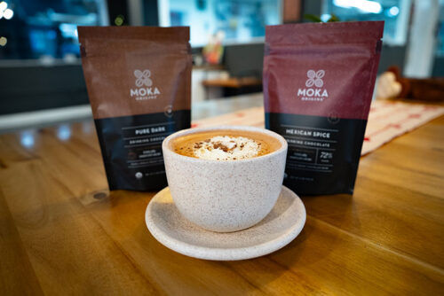 A mug of hot chocolate topped with whipped cream sits on a table besides two bags of coffee from Moka Origins in Honesdale, PA.